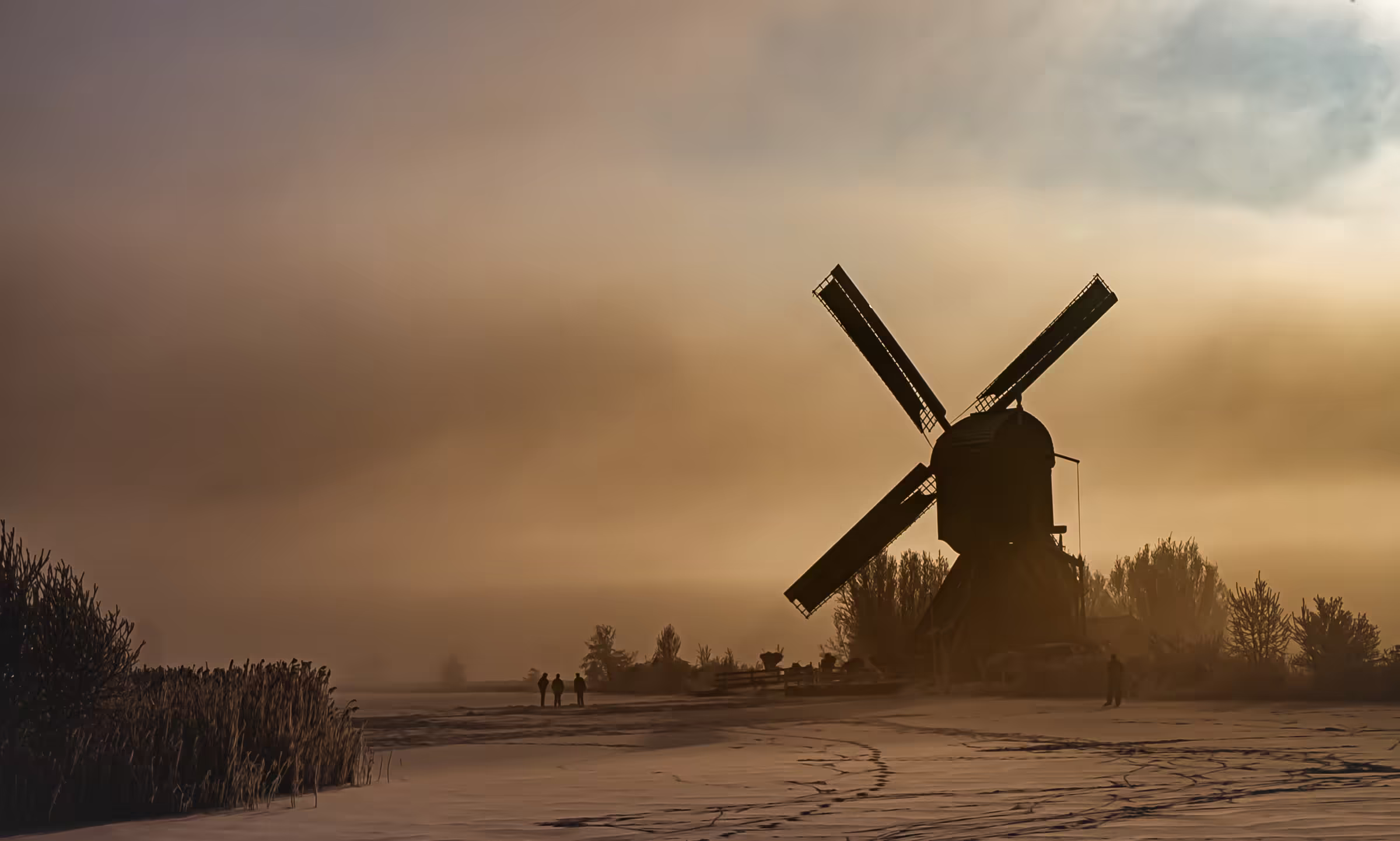 Landscape photograph of a Dutch windmill silhouetted against a misty, golden sky. The windmill's dark form stands prominently on a snowy or frosty field.